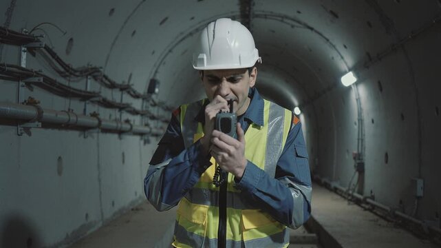 Male worker in hard hat speaks into walkie talkie inside concrete tunnel. Scene conveys urgency or coordination in industrial setting. Perfect for construction and engineering projects.