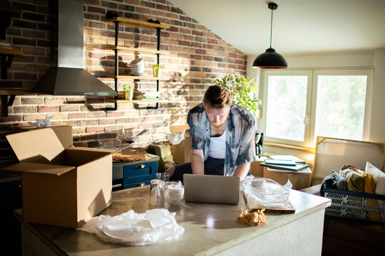 Young man using laptop amid moving boxes in modern kitchen