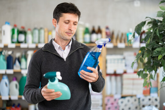 Dark-haired man stands near store counter and chooses kitchen cleaner spray. Buyer examines packaging and selects, buys household chemistry. Client see over good in household shop