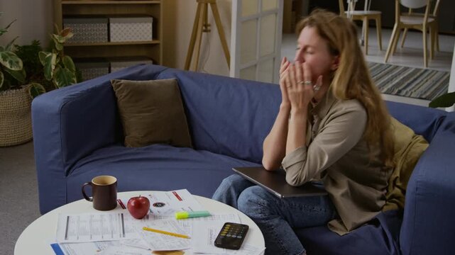 High angle shot of tired woman using laptop and rubbing eyes while sitting on couch at home and working with documents scattered on table