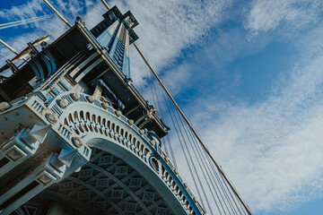 View of the Manhattan Bridge structure under blue sky with clouds. The view captures the underbelly of the Manhattan Bridge with cables and stone details against the sky.