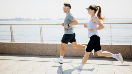 Two people run along a waterfront path on a sunny day near a city skyline with water in view