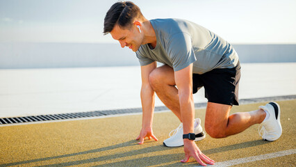 Man prepares to run on a track under clear sky during morning or afternoon with earbuds and sports...