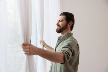 Man opening stylish window curtains at home