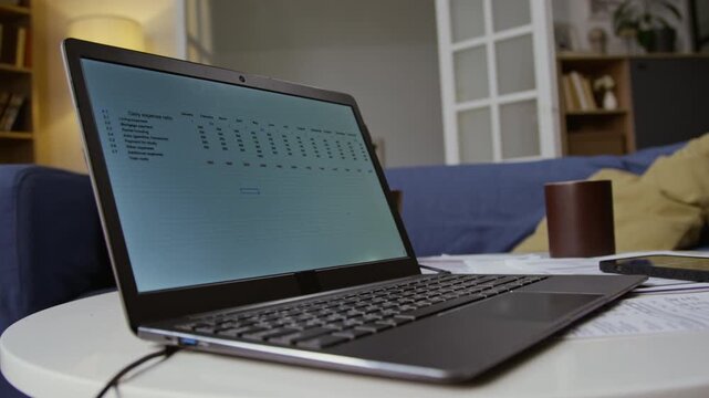At angle close up shot of open laptop with budgeting data on screen set on coffee table with scattered financial documents, copy space
