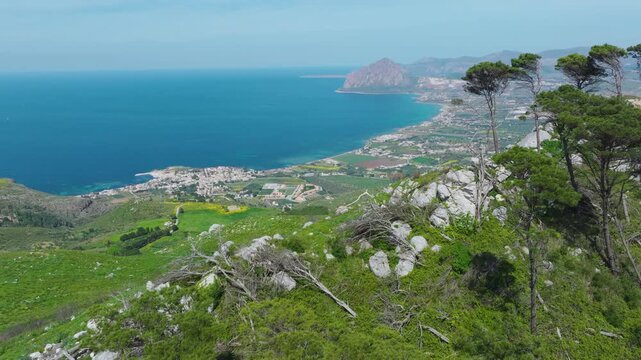 AERIAL Sweeping view of Trapani with lush green hillsides and rocky outcrops. Seaside town and mosaic of farmland stretch along coast under blue sky. Picturesque views while exploring island of Sicily