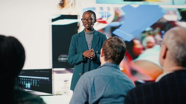 African American female speaker delivers powerful media presentation at tech conference, diverse audience in modern conference hall. Young adult discusses advertising and public relations. Camera B.