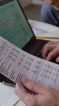 Vertical closeup shot of male hand holding financial documents while filling in online tax forms via computer while couple planning budget together