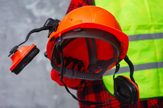 Orange safety helmet with hearing protection earmuffs is held in the hand of a construction worker wearing a yellow vest. Personal protective equipment on industrial and construction sites.