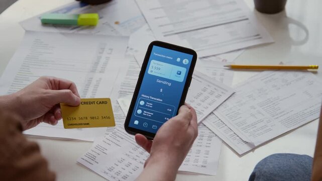High angle close up shot of male hands holding credit card and phone with online banking app on screen and money transfer transaction in progress over documents scattered on table, copy space