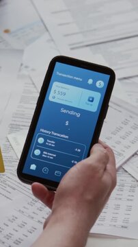Vertical closeup shot of male hands holding credit card and smartphone with online banking app on screen and money transfer transaction in progress over documents scattered on table