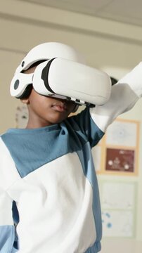 Vertical video: Reaching kid in blue-white sweatshirt using VR headset, extending arm in classroom