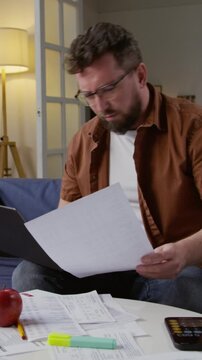 Vertical shot of adult man using computer while sitting on sofa at home and working with finances various documents scattered on table