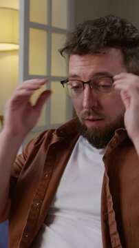 Vertical shot of exhausted adult man closing computer and rubbing eyes while sitting on sofa working from home various documents scattered on table