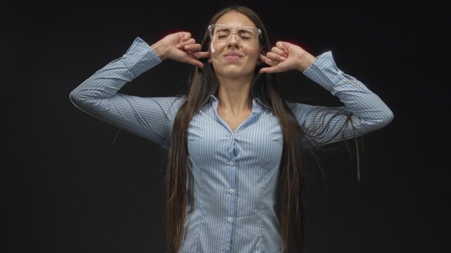 Teenage woman plugs ears with fingers in a studio with black backdrop and long hair visible; sensory overload frustration.