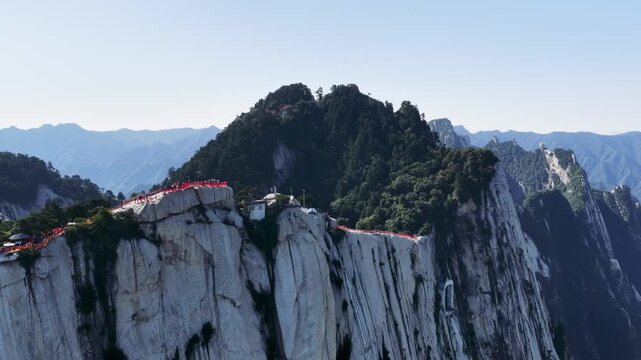 Aerial footage of the West Peak at Mount Hua, dramatic cliffs, forested ridges, and a cable car system, tourists walking along narrow paths on sheer rock faces. Shaanxi Province, China.