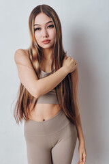 Fototapeta premium Young woman with long straight hair wearing beige sportswear posing indoors against white background. Fitness outfit, natural makeup and casual confident expression for lifestyle.