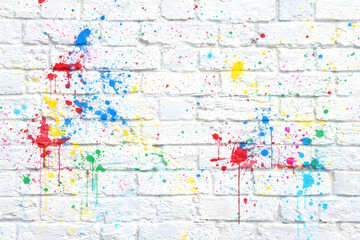 Abstract background of a white brick wall texture heavily splattered with bright, colorful paint drops, splashes, and drips.