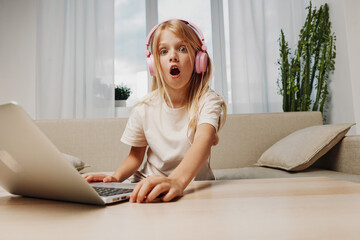Obraz premium Surprised girl with headphones uses laptop at home, sitting on couch in bright room. Concept of online learning, digital communication, and children's curiosity.
