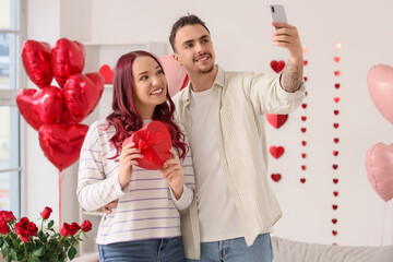 Young lovely couple with gift and heart-shaped balloons taking selfie at home. Valentine's Day...