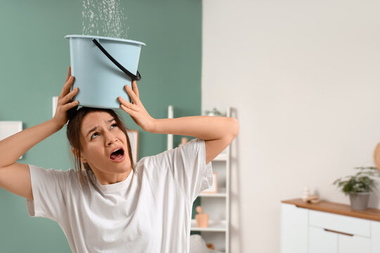 Shocked young woman placing bucket under water dripping from ceiling in room