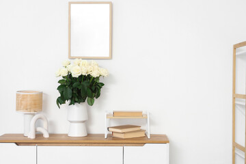 Fototapeta premium Vase with white roses, books and lamp on chest of drawers in living room