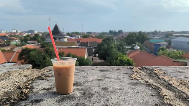 A plastic cup of iced coffee with a red straw placed on a concrete rooftop, overlooking a quiet urban neighborhood under a bright blue sky.