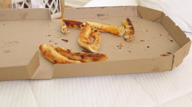 An open and nearly empty brown cardboard pizza box containing leftover pizza crusts is placed on a white bed in a softly lit room with a basket visible in the background.