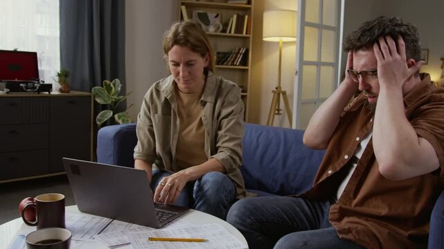 Medium shot of troubled adult couple managing finances at home with man sighing deeply and holding head in foreground trying to figure out numbers and budgeting