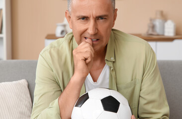 Worried mature man with soccer ball biting nails while watching TV at home