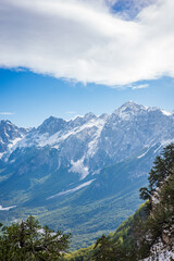 Fototapeta premium A breathtaking alpine panorama showcasing towering snow covered peaks rising above a deep green forested valley. Soft clouds drift across a bright blue sky