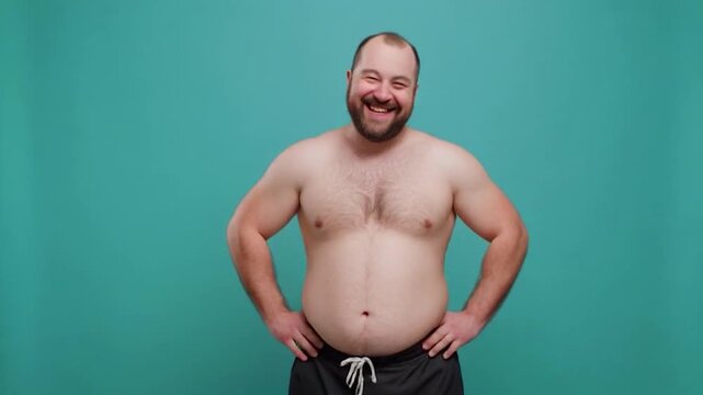 Smiling man with a beard poses shirtless in front of a teal background.