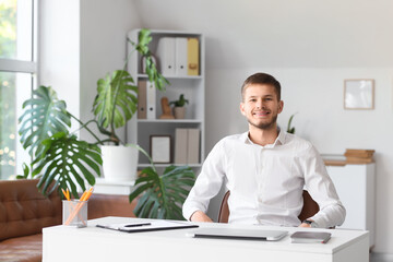 Fototapeta premium Handsome young happy businessman with laptop sitting at table in office