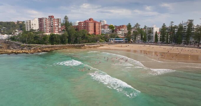 Panoramic aerial view people swimming in the turquoise Pacific Ocean water at Manly Beach, with sandy shores, bustling promenade, and coastal buildings lining the popular Sydney suburb. Drone panorama