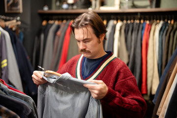 Mustached man shopping for shirts in second hand store, choosing clothing, inspecting size and...