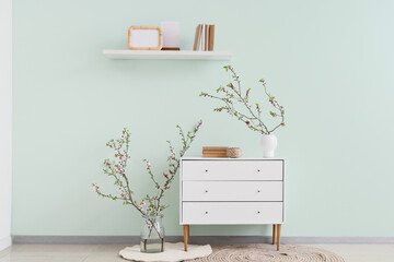 Vases with blooming branches, chest of drawers and shelf near green wall © Pixel-Shot