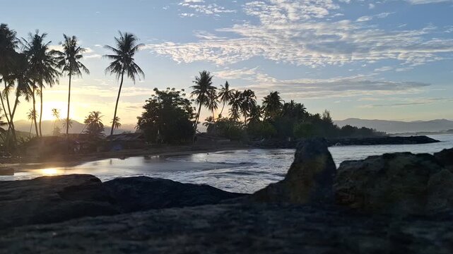 Hyperlapse sunrise over coastal village with sunlight through coconut trees Cocos nucifera and Bukit Barisan mountains, golden hour light moving over houses near beach, tropical landscape time-lapse