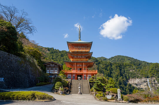 Striking three-story pagoda stands at the foot of lush green hills, with a distant waterfall cascading down rocky cliffs under a clear blue sky at Kumano Nachi Taisha, Wakayama, Japan. Bright sunlight
