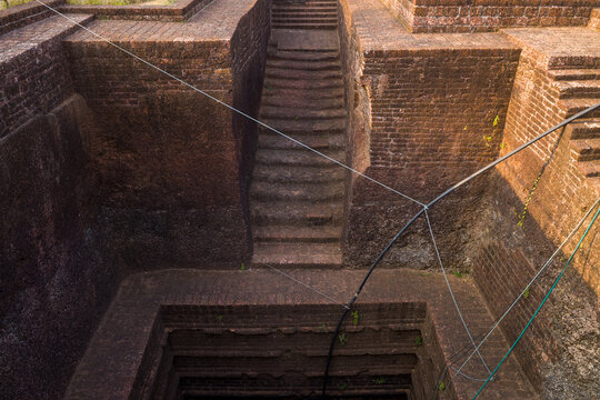Symmetrical stone steps descend into a deep, geometric stepwell surrounded by weathered brick walls at Bekal Fort, India. Warm afternoon light highlights the textured surfaces and architectural