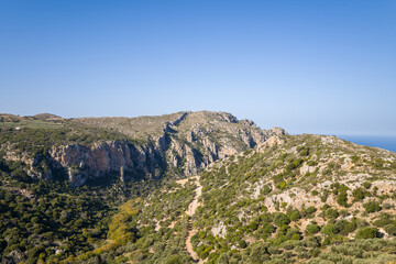 Fototapeta premium Expansive view of a rugged mountain ridge in western Crete, featuring rocky cliffs, scattered shrubs, and a winding dirt trail under a clear blue sky. The landscape is sunlit and open, evoking a sense