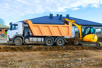 Dump truck is parked next to house while an excavator digs in dirt at construction site.