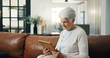 Naklejka premium Senior woman, photo frame and sad on sofa with memory, grief and nostalgia in lounge at house. Elderly patient, picture and remember with depression, mental health and broken hearth in living room