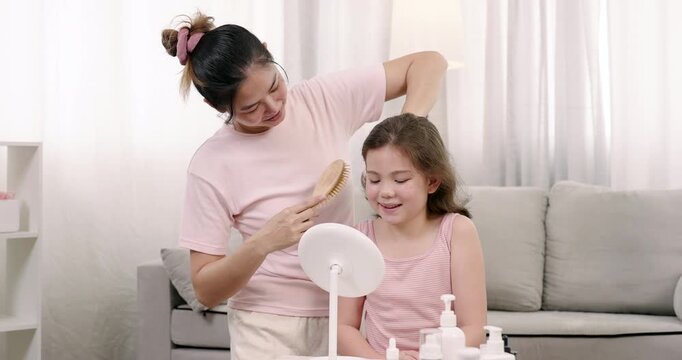 Asian adult female single mother gently pulling multiracial young girl's hair to tie while girl smiles watching mirror during slowmotion bonding time in peaceful beauty routine at home living room