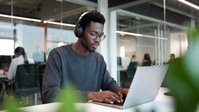 A focused young Black man wearing headphones and glasses works intently on a laptop in a modern office.