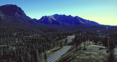 sunlit highway cutting through pine forest morning, golden glow on asphalt, long perspective toward jagged peaks, cinematic mood, ideal roadtrip backdrop