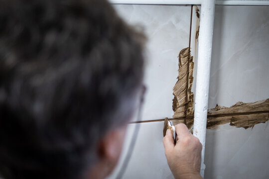 Worker applies brown grout around a vertical pipe and cross joint on glossy tiles using a small plastic tool. The renovation scene focuses on careful sealing in tight areas for a durable, waterproof
