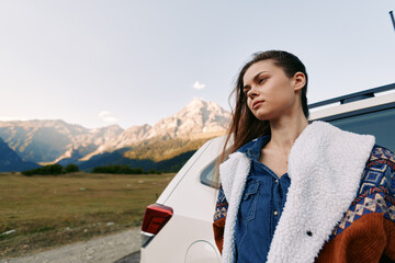 Obraz premium Woman by car in mountain landscape, travel portrait of a young woman leaning on a vehicle beside a rural road, scenic outdoor adventure with peaks, meadow and thoughtful expression