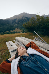 Obraz premium woman lying on a wooden bench in a meadow with mountains in the background, portrait of relaxation outdoors in countryside, casual denim jacket and shearling coat, peaceful scenic escape