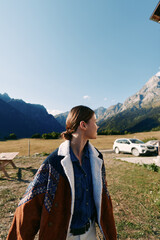 Obraz premium Woman in a warm jacket standing in a meadow by a white car, looking toward distant mountains during travel. Scenic outdoor portrait of nature, adventure and countryside road trip.