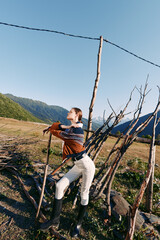 Obraz premium Woman leans on rustic fence in countryside meadow with mountains in background, wearing sweater and boots while arranging wooden sticks for rural fence repair and outdoor work.
